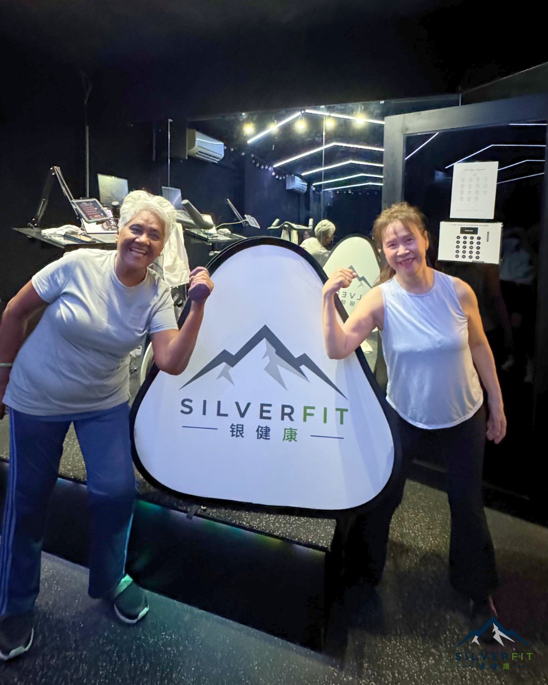 Two SilverFit seniors smiling together beside the SilverFit branded sign in the gym
